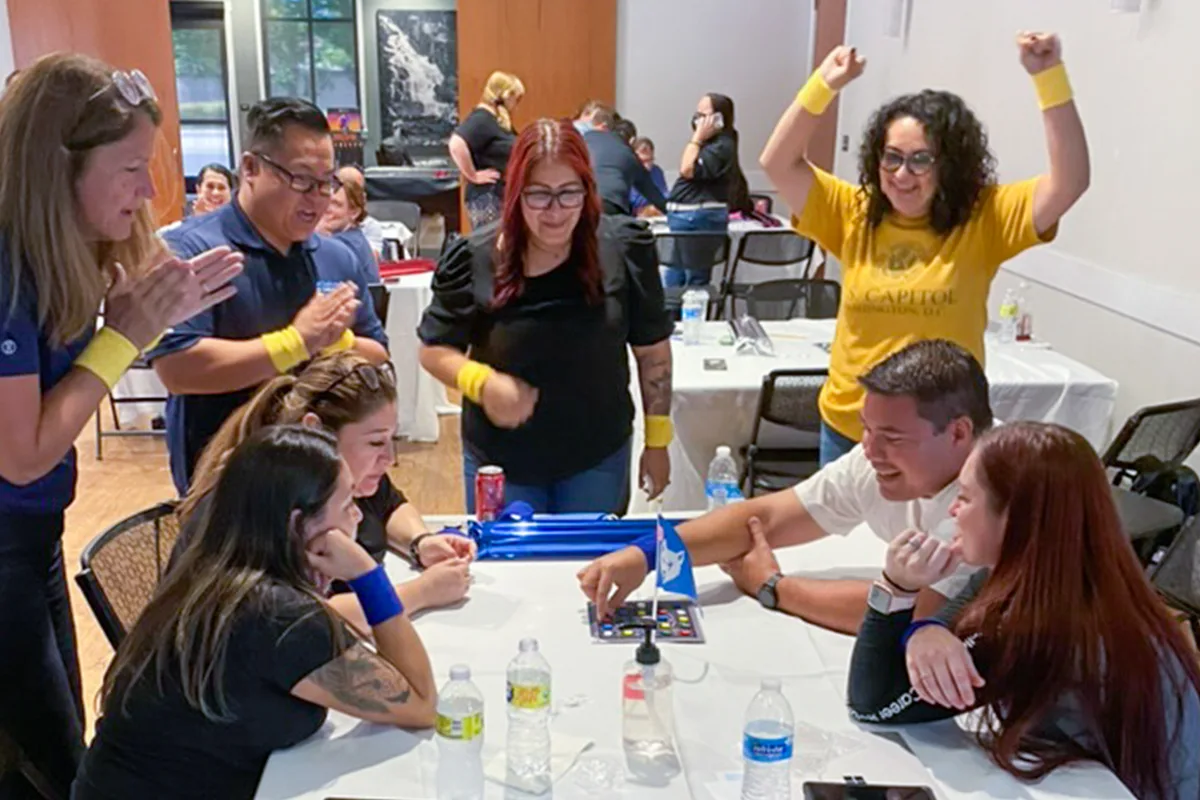Educators in matching wristbands cheer around a table as a team plays an UNLOCK game board